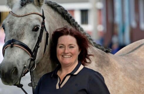 Woman with a black and cream top standing beside a large grey horse with a braided mane