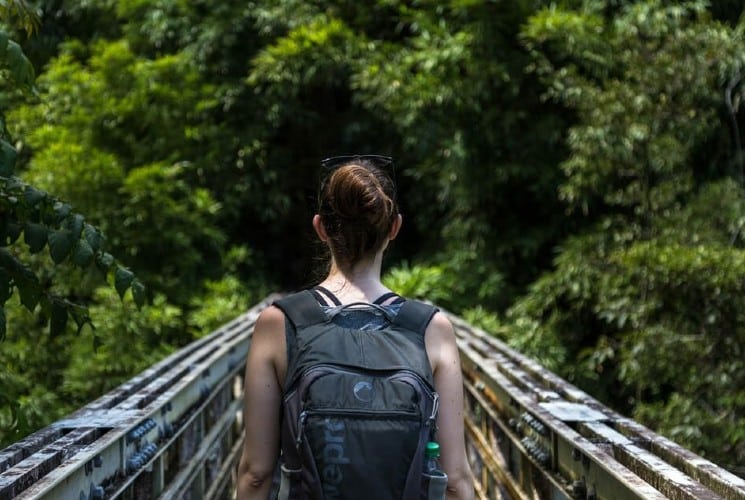 A woman wearing a backpack walking acrpss a bridge surrounded by trees