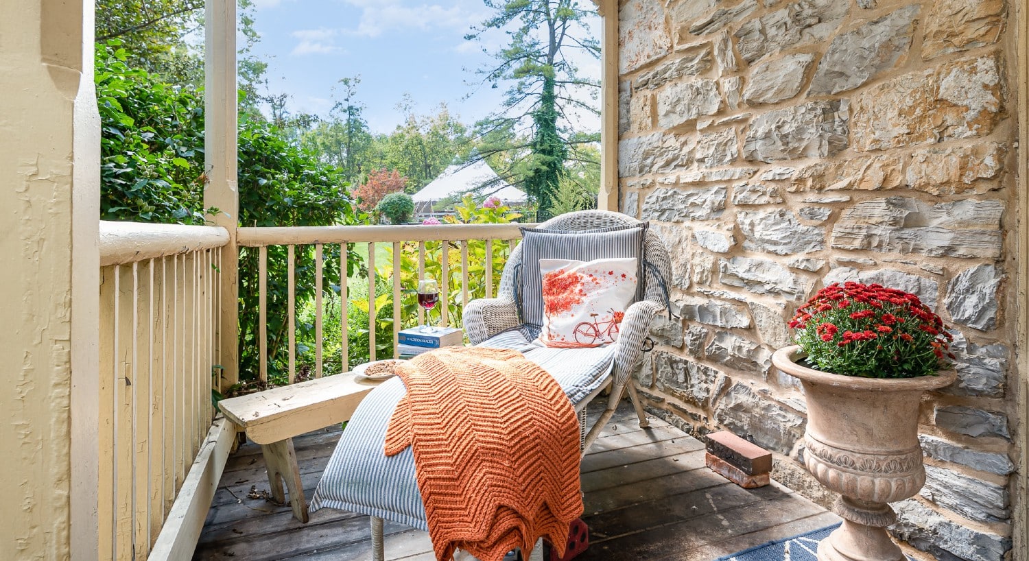 A wicker chaise lounge chair with pillow and orange blanket at the corner of an outdoor patio overlooking trees and a blue sky
