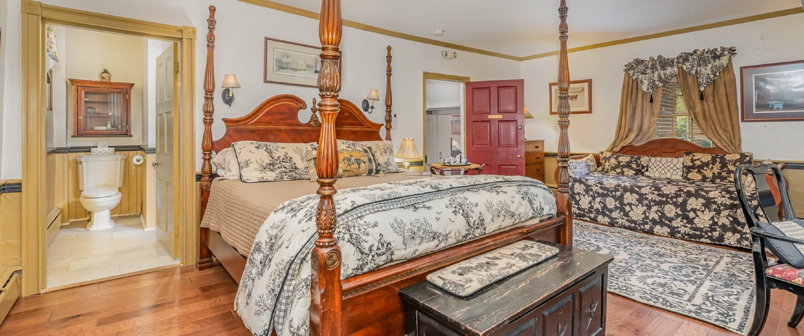 Large bedroom with four-poster bed, floral couch, hardwood floors and doorway into a bathroom