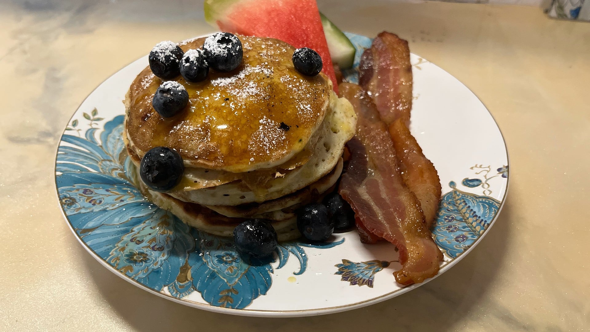 Delicious stack of blueberry pancakes with watermelon and bacon on a white and blue floral plate