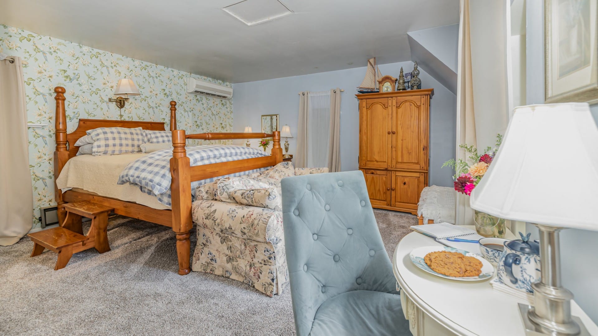 Cozy and elegant bedroom in blue hues with wood bed, floral couch, armoire and desk with sitting chair