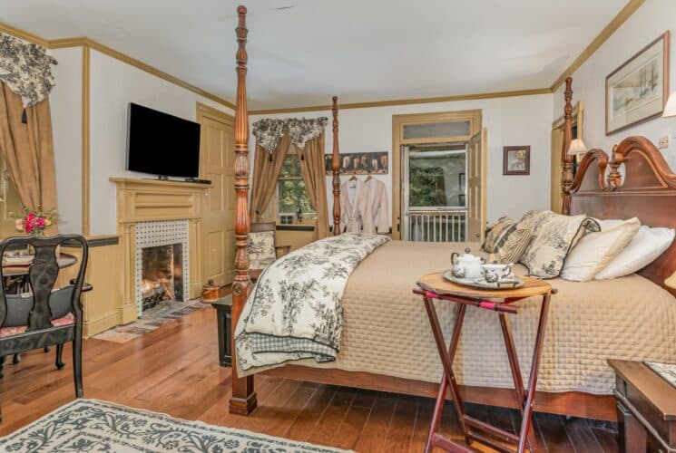 Bedroom with four-poster bed, large windows with curtains, table for two and TV over a fireplace