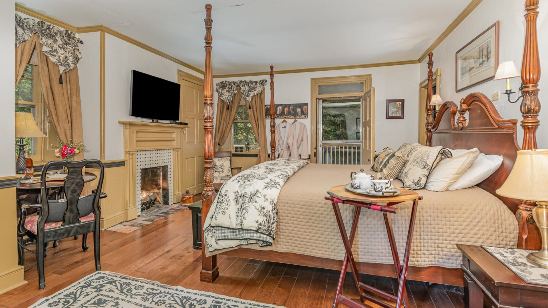 Bedroom with four-poster bed, large windows with curtains, table for two and TV over a fireplace