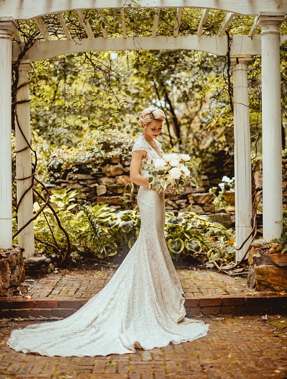 Stunning bride in a wedding gown with bouquet standing under a white pergola surrounded by trees