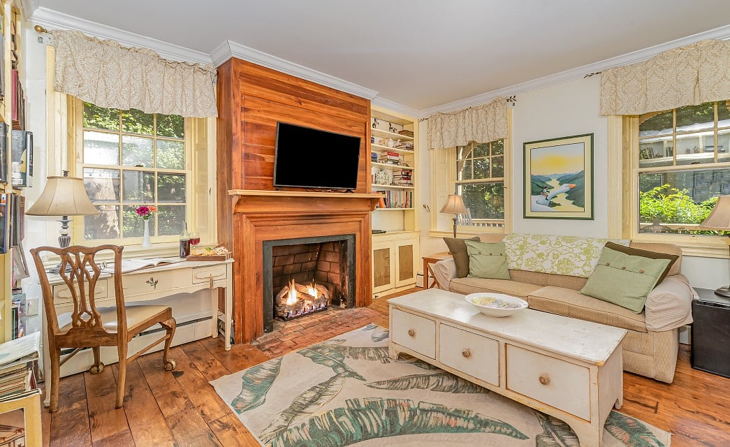 Bright living room area wit couch, coffee table, writing desk, large windows and TV over a fireplace