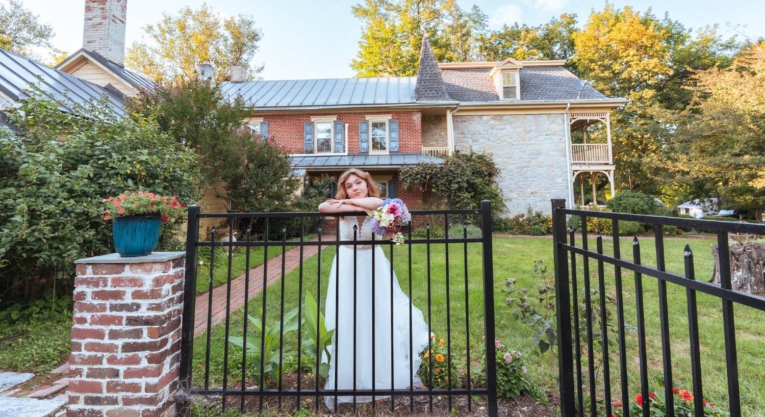 A bride in white with a colorful bouquet standing by a black iron fence in front of a large brick home