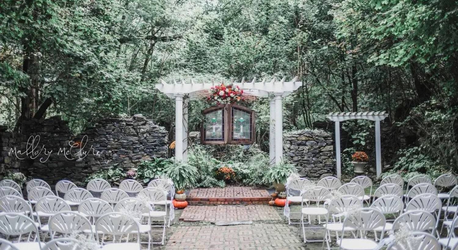 A gorgeous white pergola and chairs set up for a wedding, set among towering trees and green landscaping