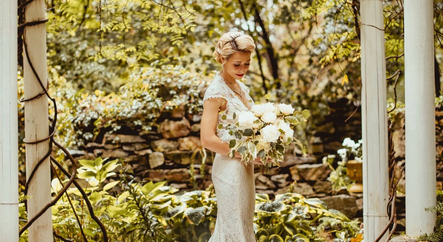 Stunning bride in a wedding gown with bouquet standing under a white pergola surrounded by trees