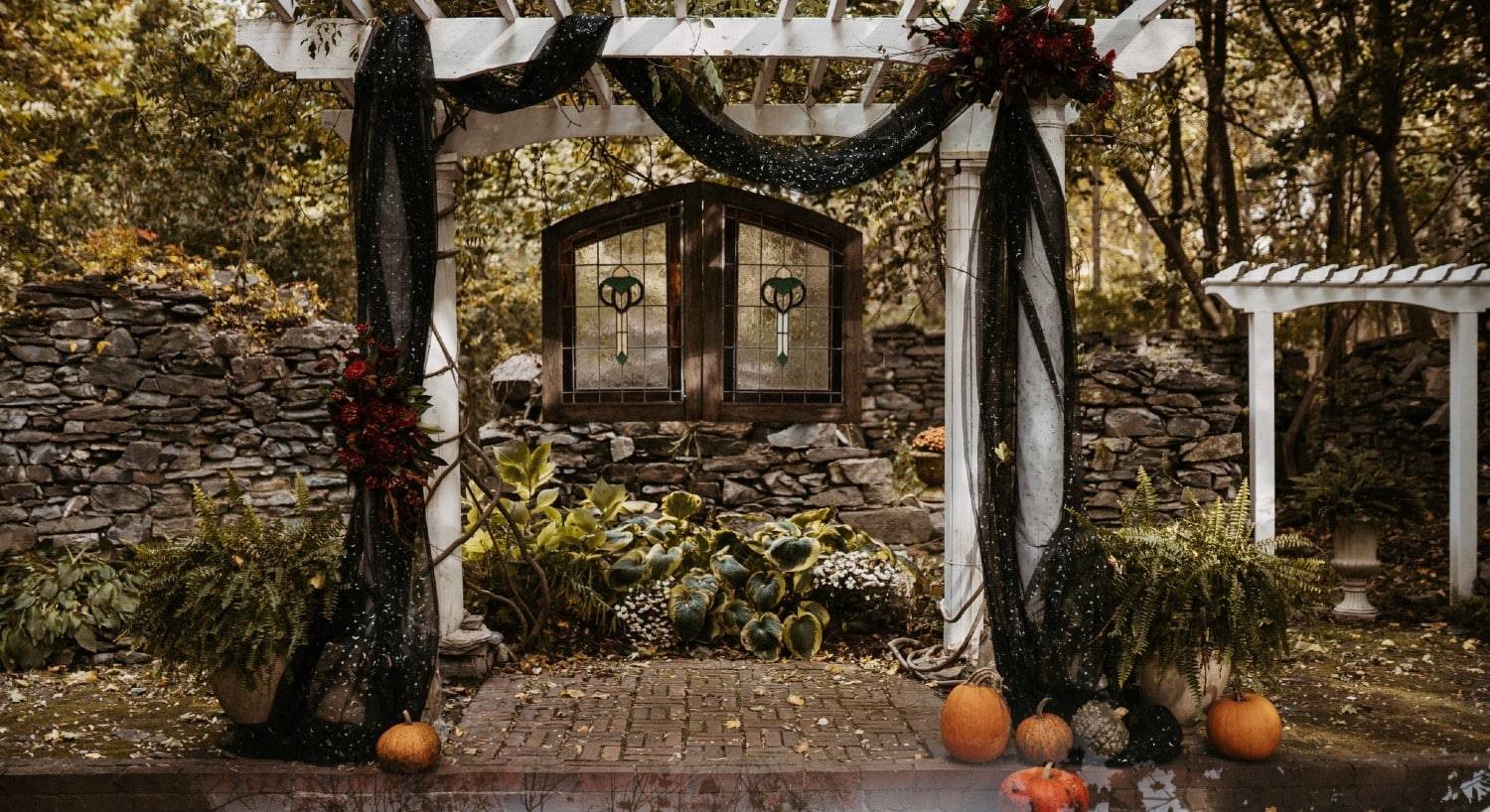 A gorgeous white pergola decorated for a fall wedding against a large stone wall surrounded by trees
