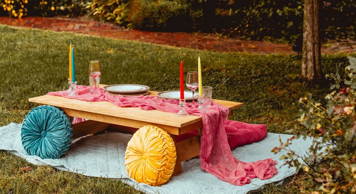A small outdoor table for two set on a blanket on the grass with colorful pillows