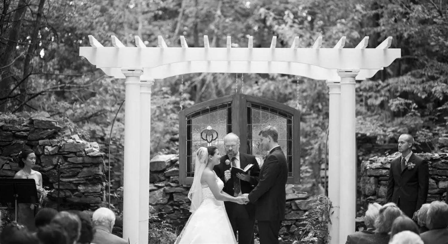 A bride and groom holding hands in front of an officiant under a white pergola outdoors