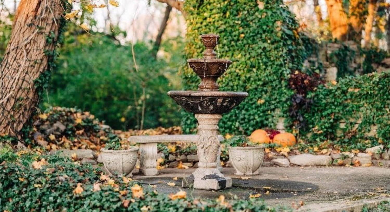 A three-tiered stone fountain next to a stone bench surrounded by lush green trees