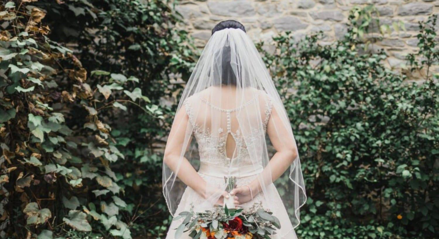 A bride in a white gown and veil holding her bouquet behind her back while facing a wall full of green bushes