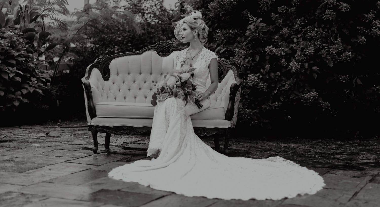 Black and white photograph of a bride in white with a bouquet sitting on an antique loveseat outdoors
