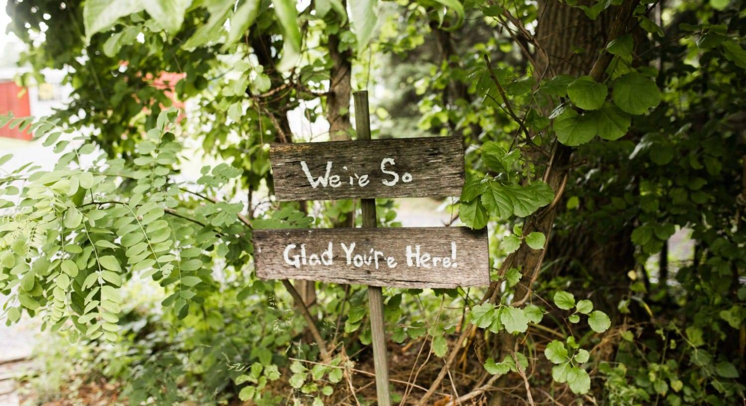 A rustic wooden sign in the woods with words "We're so glad you're here!"