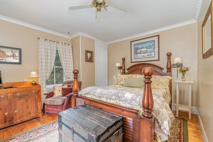 Bedroom with four-poster bed, chest, wood hutch, velvet chair and windows with plaid curtains