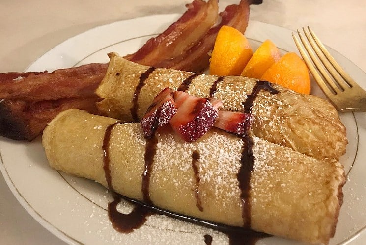 Breakfast place of two crepes drizzled with chocolate with sides of fruit and bacon