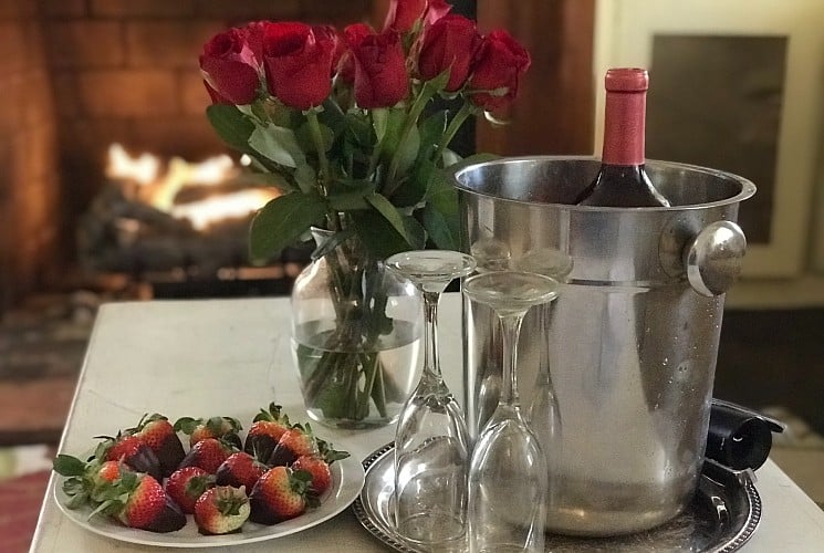 Plate of chocolate covered strawberries next to a vase of roses and silver bucket with champagne and two glasses