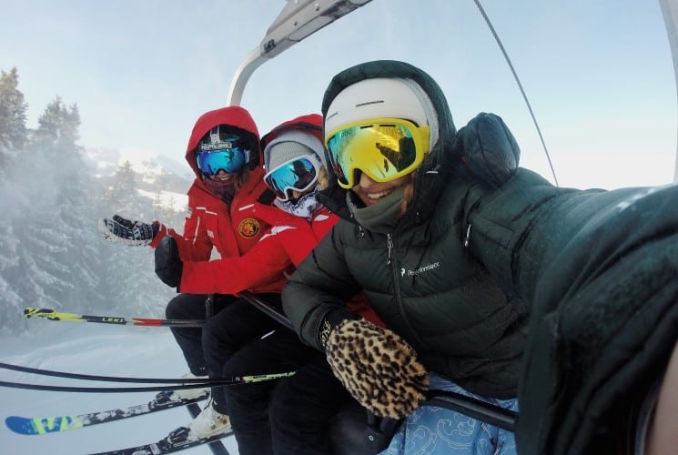 Three people taking a selfie while on a ski chairlift surrounded by snow-covered trees