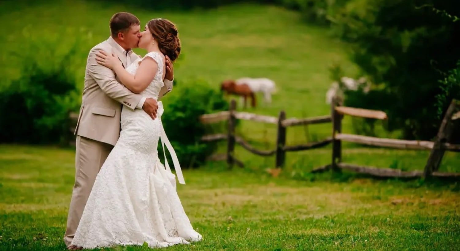 A bride and groom kissing with a fence and horse pasture in the background