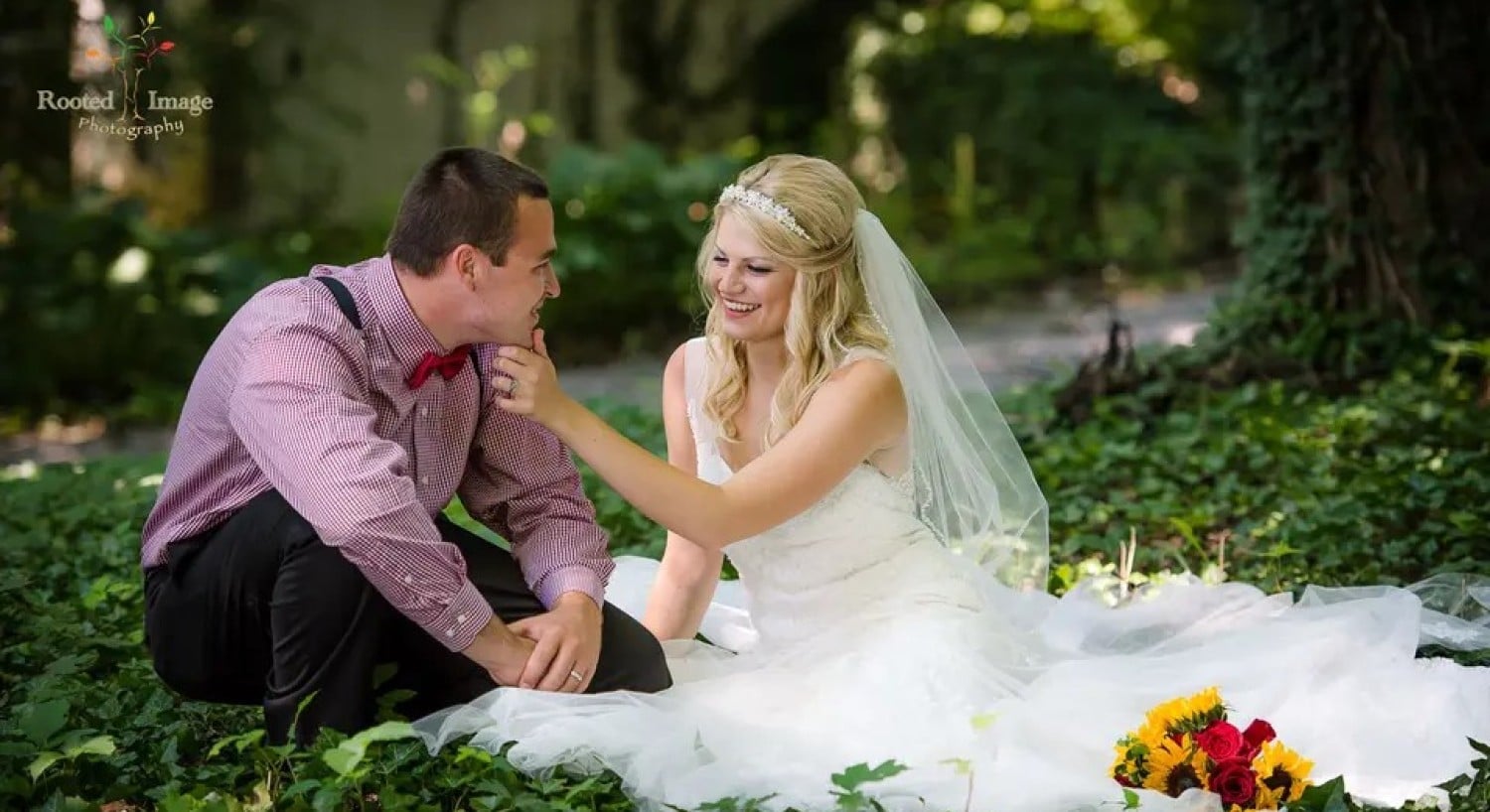 A bride and groom sitting on the ground together at the base of a tree surrounded by lush green leaves