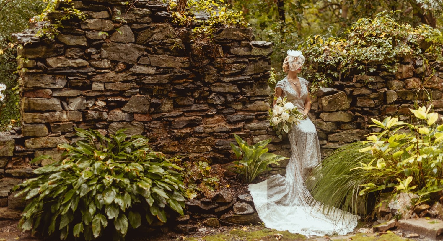A stunning bride in a white gown sitting atop a small nook in a wall of stone surrounded by trees and plants