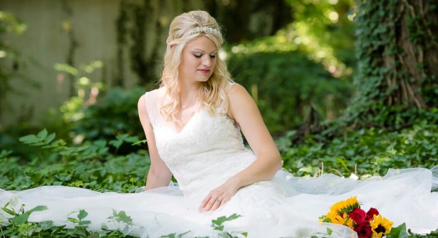 A bride in white with a red and yellow bouquet sitting at the base of three surrounded by green leaves